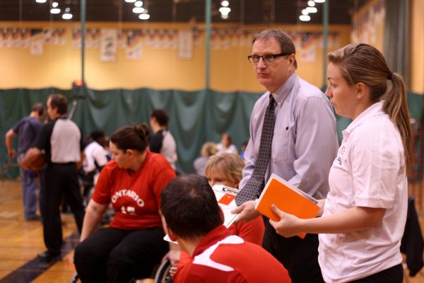 male wheelchair basketball coach wearing tie holding clipboard with female assistant coach talking to athletes at half-time