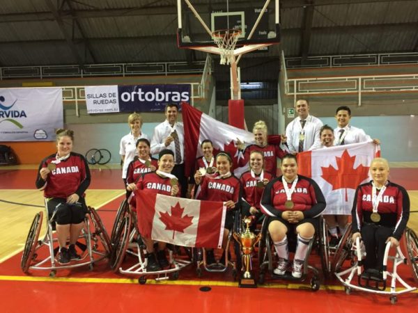 members of the Canadian Women's Wheelchair Basketball Team pose for a photo on the court while holding the tournament trophy and Canadian flags